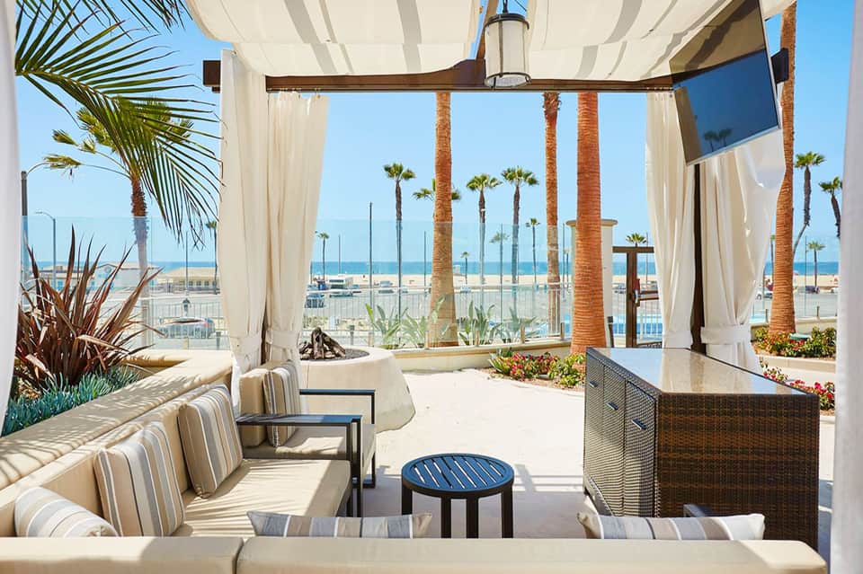 Beachfront lounge with white curtains, striped seating, and palm trees overlooking sandy beach