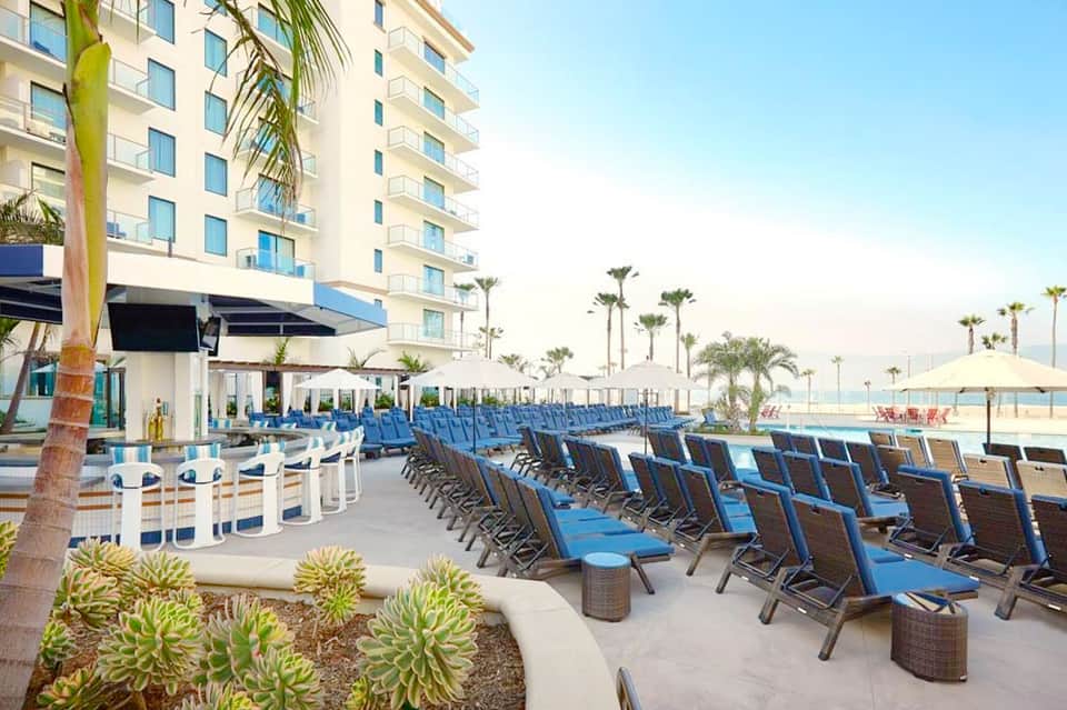 Beachfront resort with rows of blue lounge chairs on sandy patio, high-rise hotel building, and palm trees