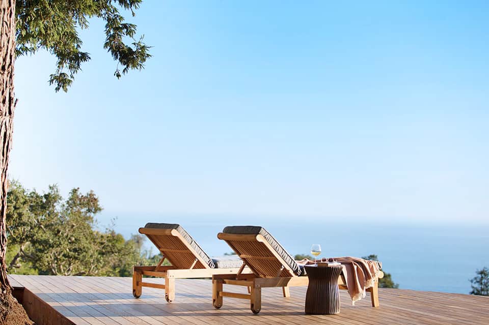 Wooden lounge chairs on oceanview terrace beneath tall trees with clear blue sky and sea horizon