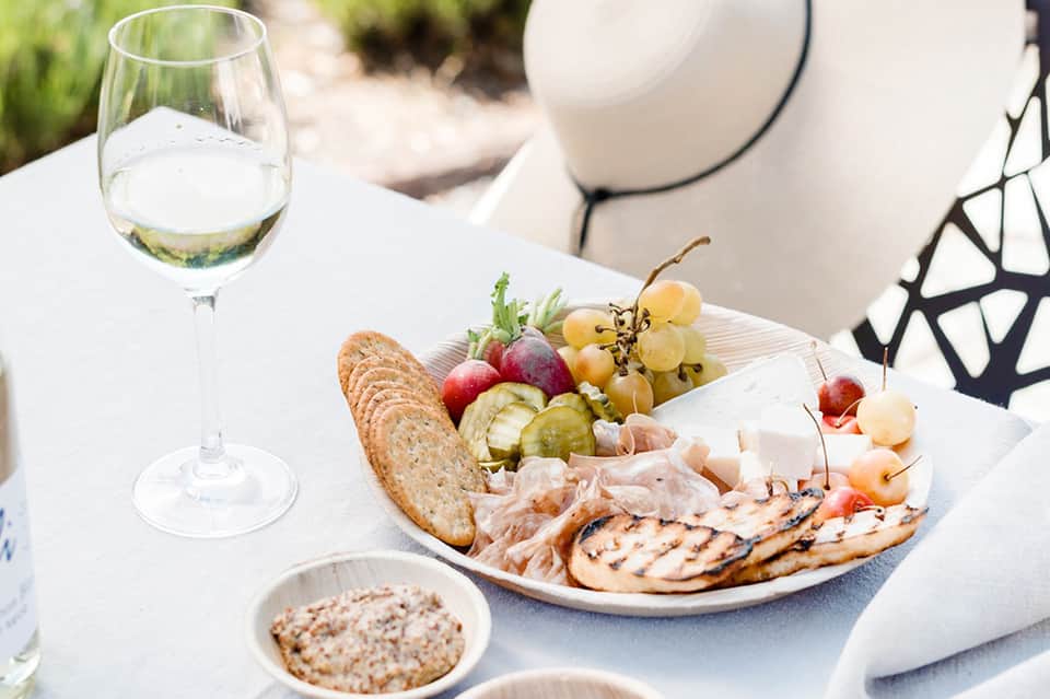 White wine glass and charcuterie board with meats, cheese, and grapes on white table