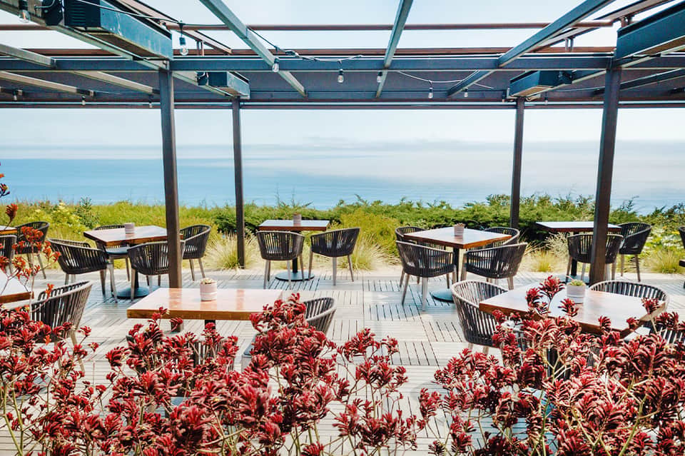 Oceanfront dining terrace with black chairs, pergola shade structure, and coastal landscape views