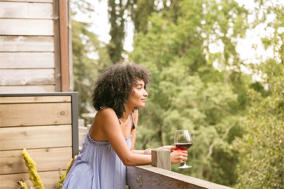 Woman in light blue dress holding red wine glass on wooden balcony surrounded by forest