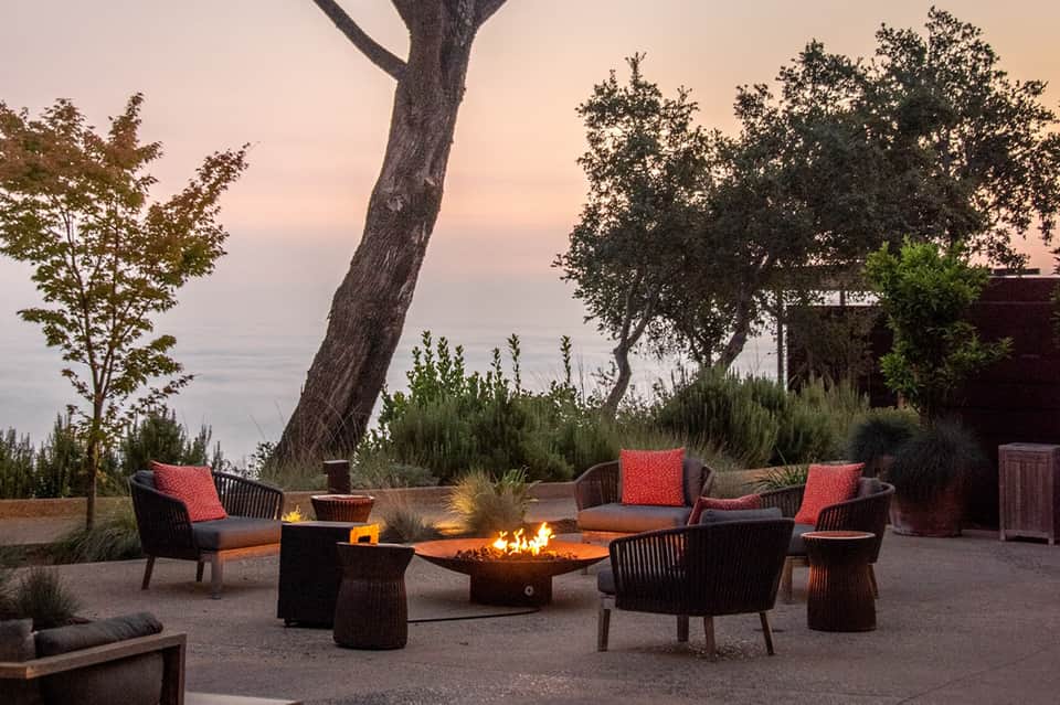 Waterfront patio with fire pit, lounge seating with red cushions, trees, and sunset ocean views