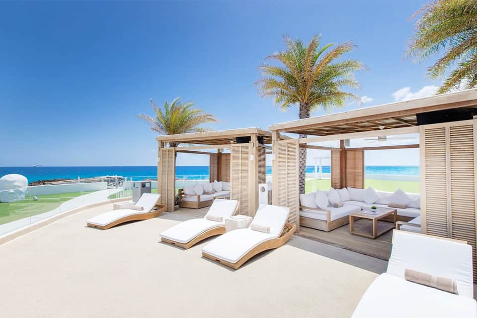 Oceanfront terrace with wooden loungers under pergolas, palm trees, and blue water beyond