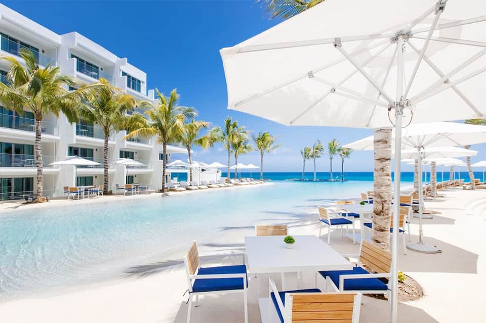 Modern infinity pool with white umbrellas and lounge chairs facing pristine white sand beach and ocean