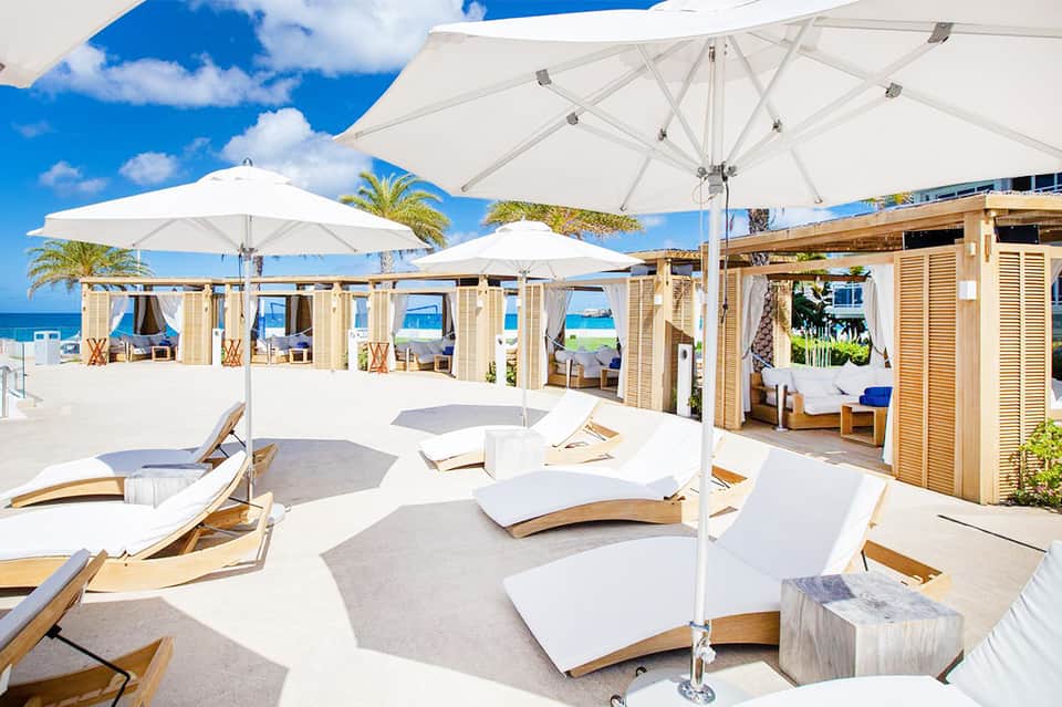 Beachfront resort with curved lounge chairs, white umbrellas, wooden cabanas, and palm trees