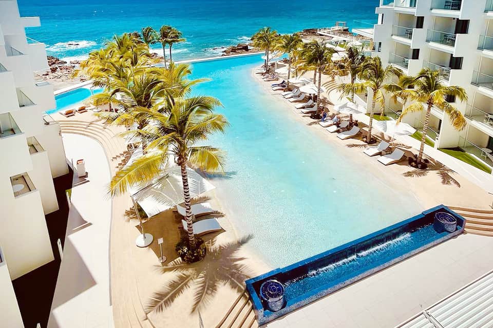 Beachfront resort pool overlooking turquoise ocean with white cabanas and palm trees