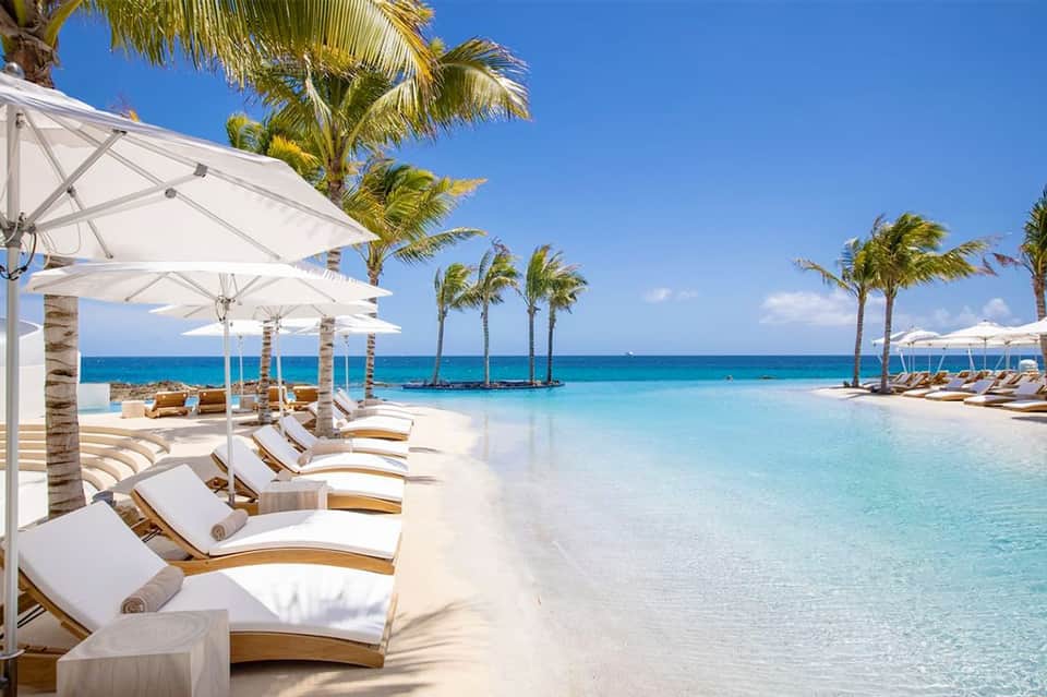 Beachfront infinity pool with white loungers and umbrellas facing turquoise ocean and palm trees