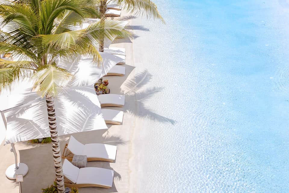 Overhead view of white lounge chairs with palm tree shadow on pristine beach by turquoise water