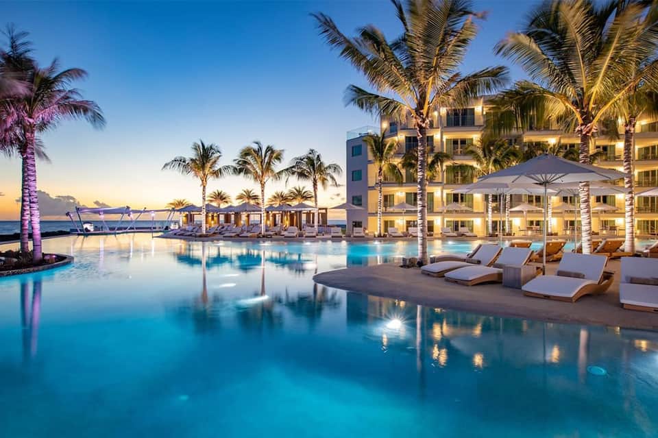 Resort infinity pool at dusk with illuminated building, palm trees, and lounge chairs