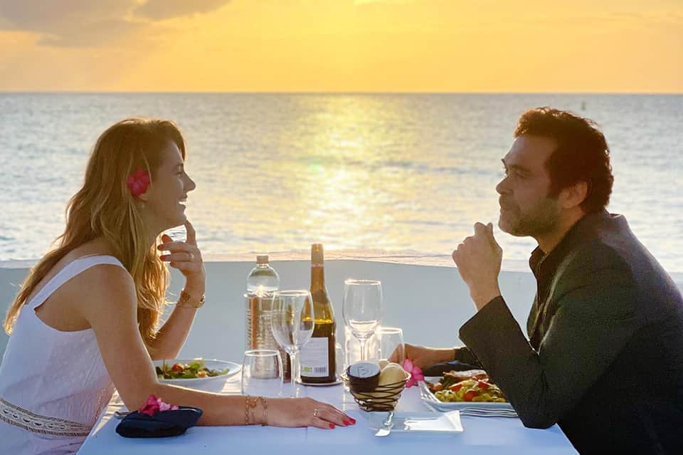Couple dining at sunset table overlooking ocean with wine, champagne, and tropical flower decorations