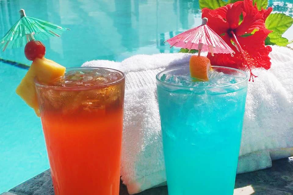 Two colorful tropical drinks poolside with umbrellas, hibiscus flower, and palm trees in background