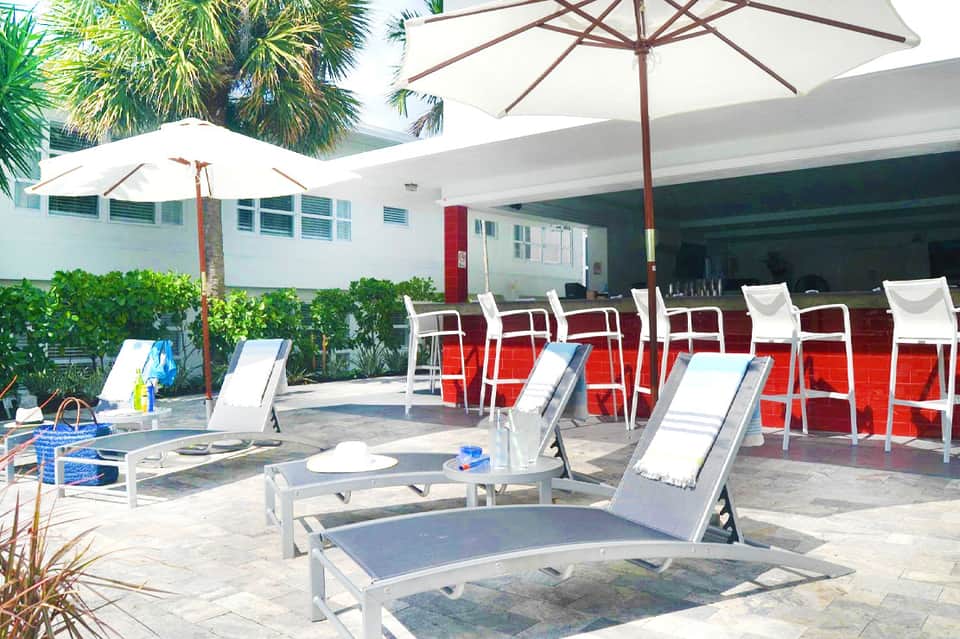 Resort pool deck with loungers, white and brown umbrellas, palm trees, and tropical landscaping