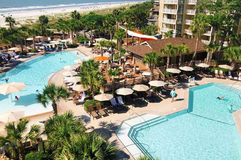 Beachfront resort with multiple pools, palm trees, and multi-story hotel building overlooking sandy beach and ocean