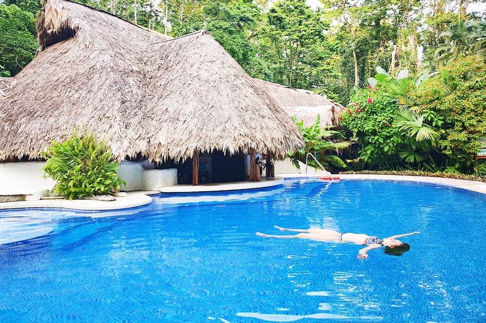 Resort swimming pool with thatched-roof cabana, tropical vegetation, and guest floating in crystal-clear blue water