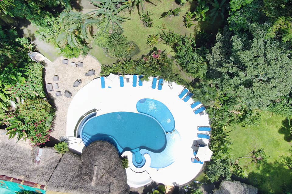 Overhead view of curved swimming pool with blue water, white deck, blue umbrellas, and surrounding tropical greenery