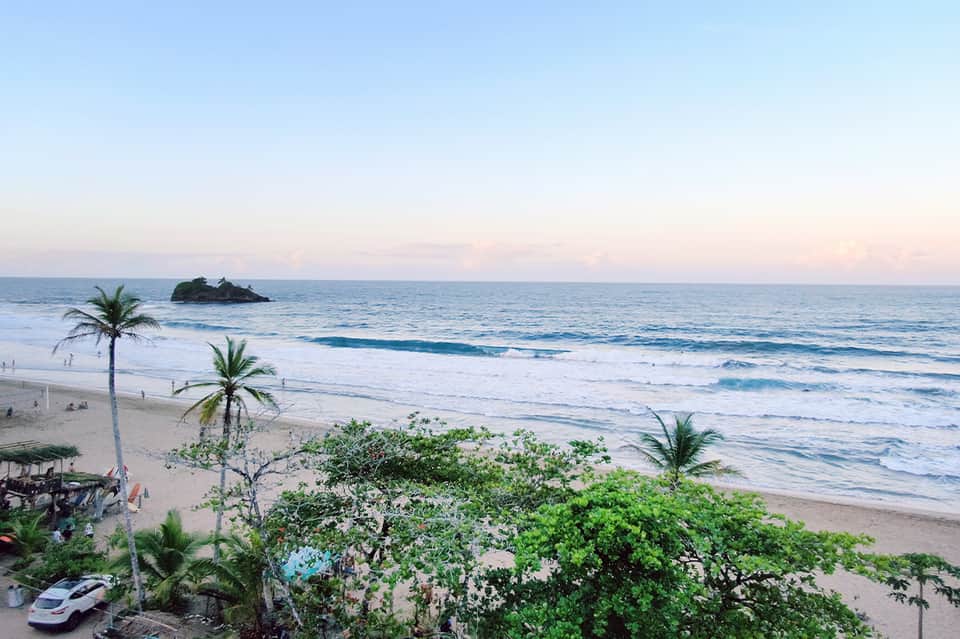 Scenic beach at dusk with palm trees, gentle waves, sandy shore, and small rock formation