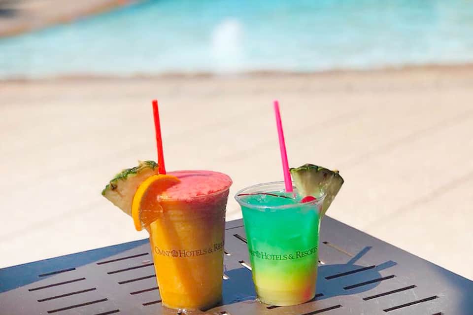 Two tropical cocktails with pineapple garnish and colorful straws on a beach table with ocean backdrop
