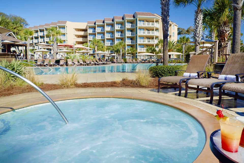 Resort pool area with multi-story beige building, lounge chairs, and palm trees on sunny day