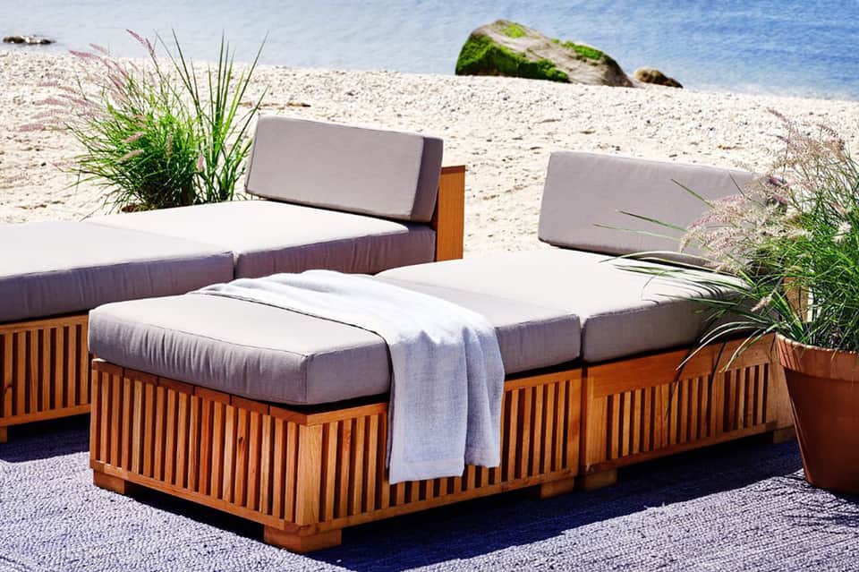 Wooden lounge chairs with gray cushions on beach next to sea grass and ocean