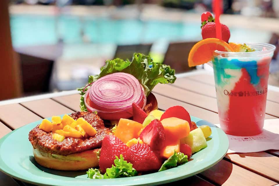Beachside brunch plate with grilled fish, fresh fruits, sliced onion, and colorful tropical cocktail by pool