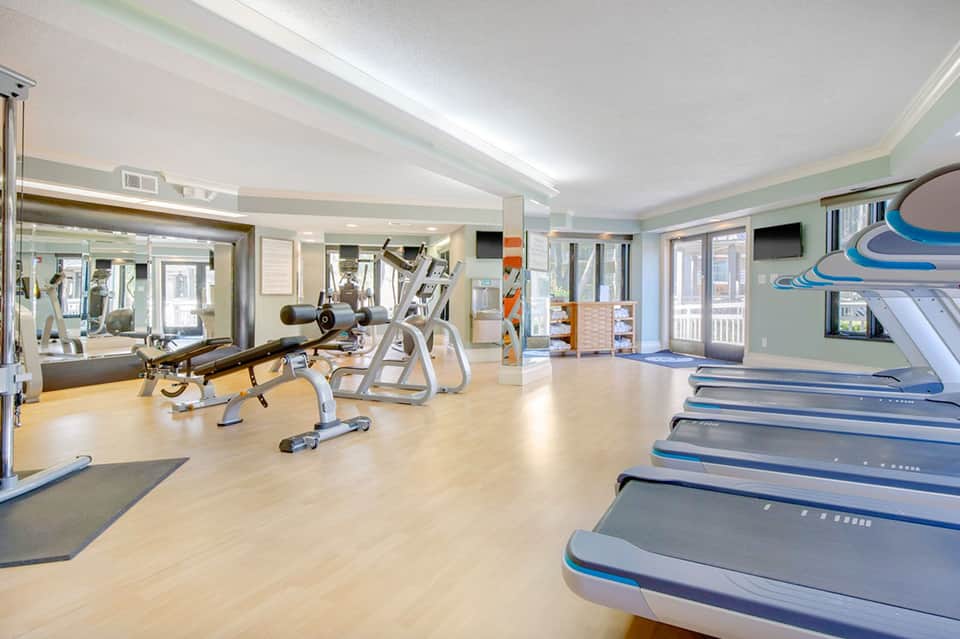 Bright fitness center with treadmills, weight machines, mirrors, and light wood flooring
