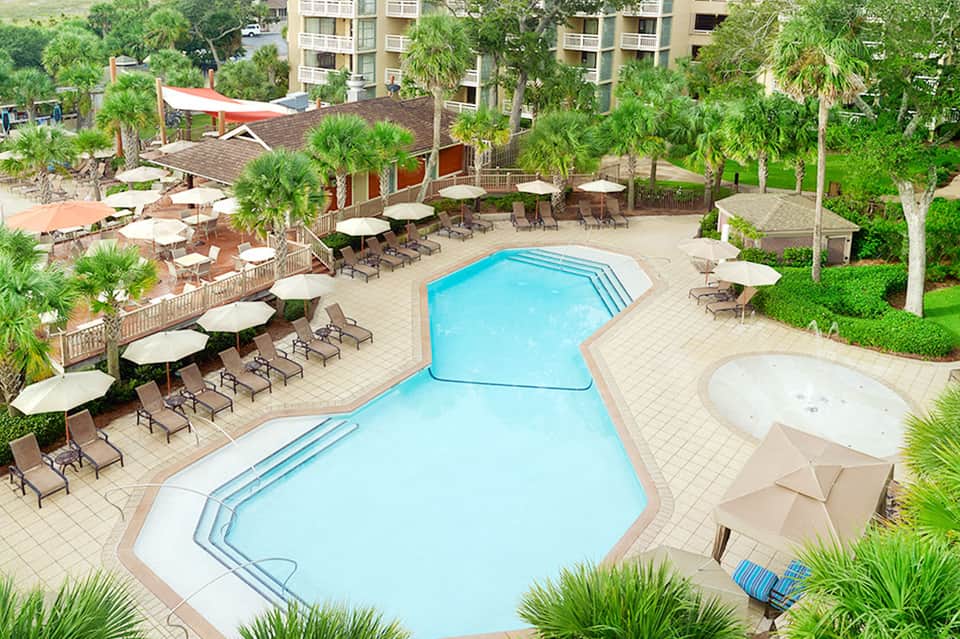 Aerial view of resort pool complex with lounge chairs, white umbrellas, palm trees, and multi-story buildings