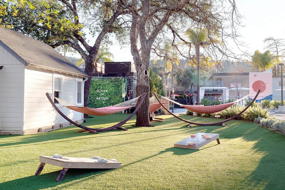 Outdoor lawn with hammocks suspended between trees, cornhole boards, and modern white buildings in background