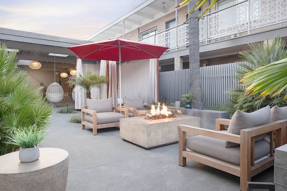 Modern courtyard with fire pit, wooden lounge chairs, red umbrella, potted plants, and contemporary architecture
