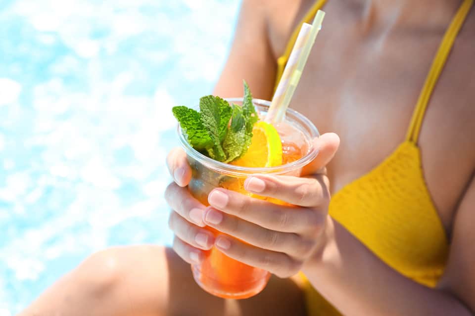 Person holding a tropical cocktail with orange juice, mint, and lemon by poolside