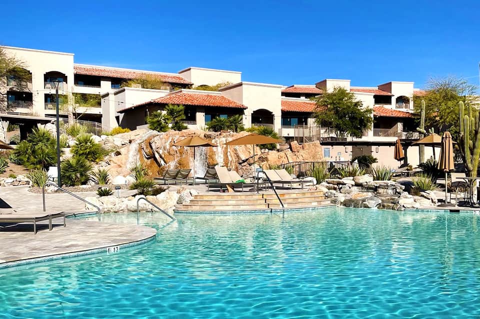 Resort pool with lounge chairs, large rocks, and white adobe-style buildings