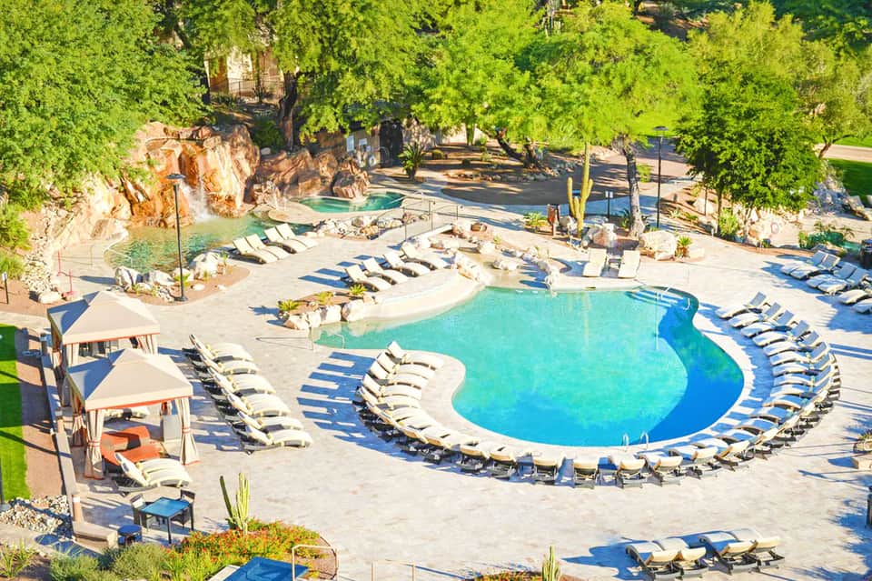 Aerial view of curved resort pool surrounded by lounge chairs, nestled among green trees and natural landscape