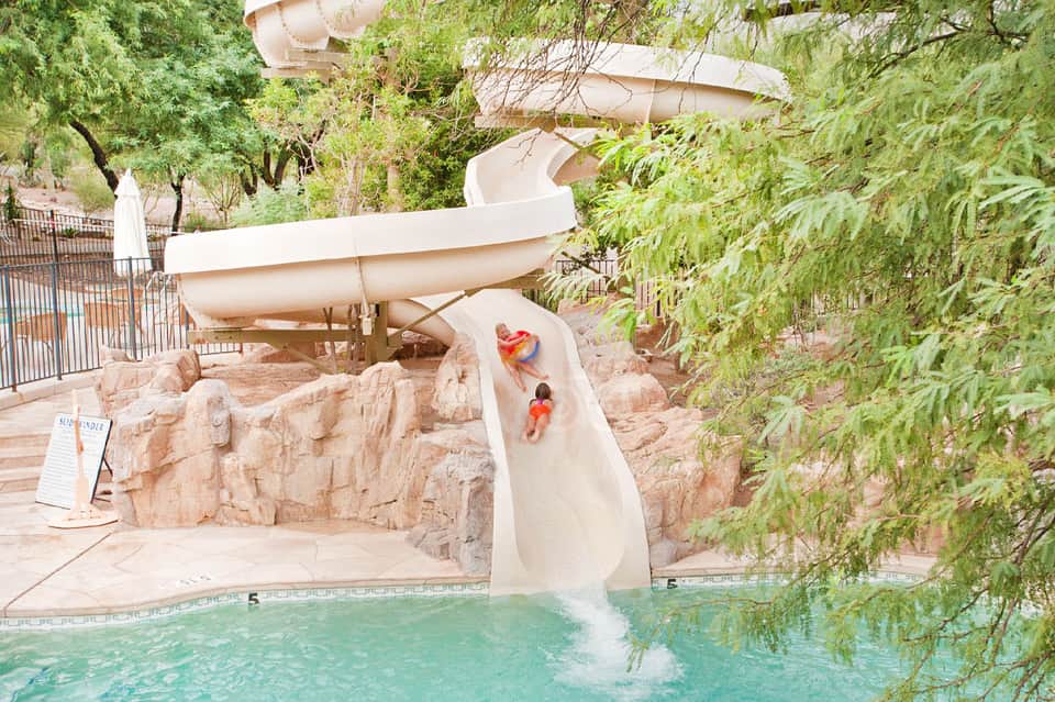 Child sliding down white waterslide into turquoise pool surrounded by natural stone landscape and greenery
