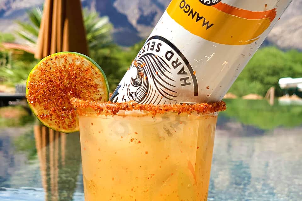 Spicy cocktail with tajín-rimmed glass and lime wheel next to bottle of hot sauce