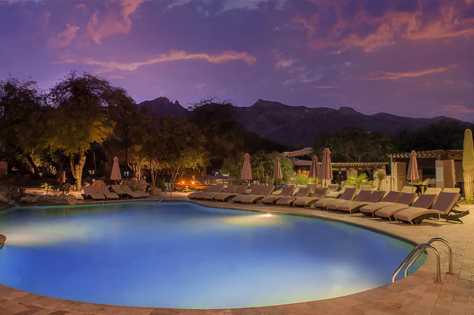Illuminated resort pool at dusk with lounge chairs, umbrellas, and mountain backdrop