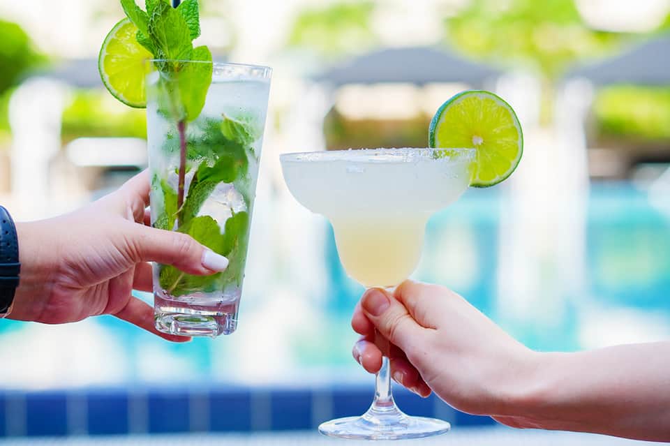 Two hands holding refreshing mojito and margarita cocktails by a swimming pool
