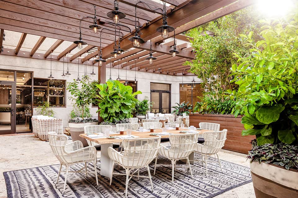 Covered patio dining area with white wicker chairs, wooden beams, pendant lighting, and lush green plants