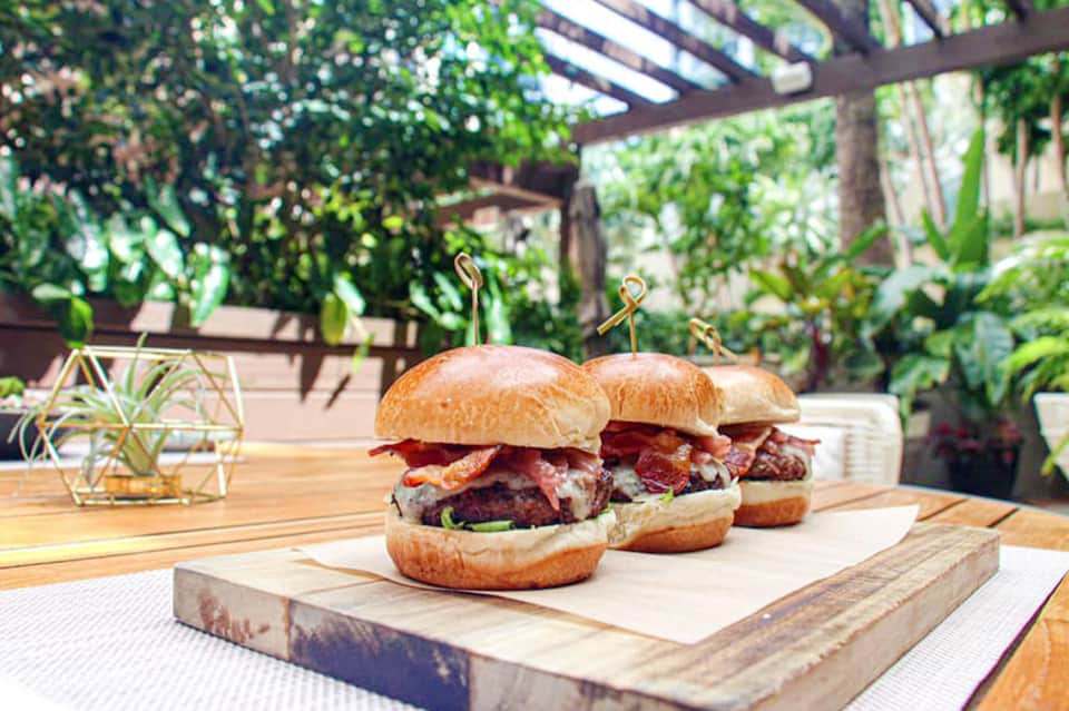 Three gourmet burgers with bacon on wooden serving board in an outdoor garden setting with greenery