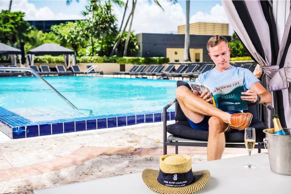 Man relaxing poolside in lounge chair reading magazine with cowboy hat on table