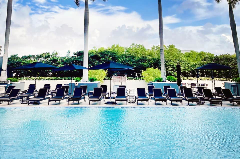 Outdoor swimming pool with lounge chairs, blue umbrellas, tall palm trees, and lush landscaping