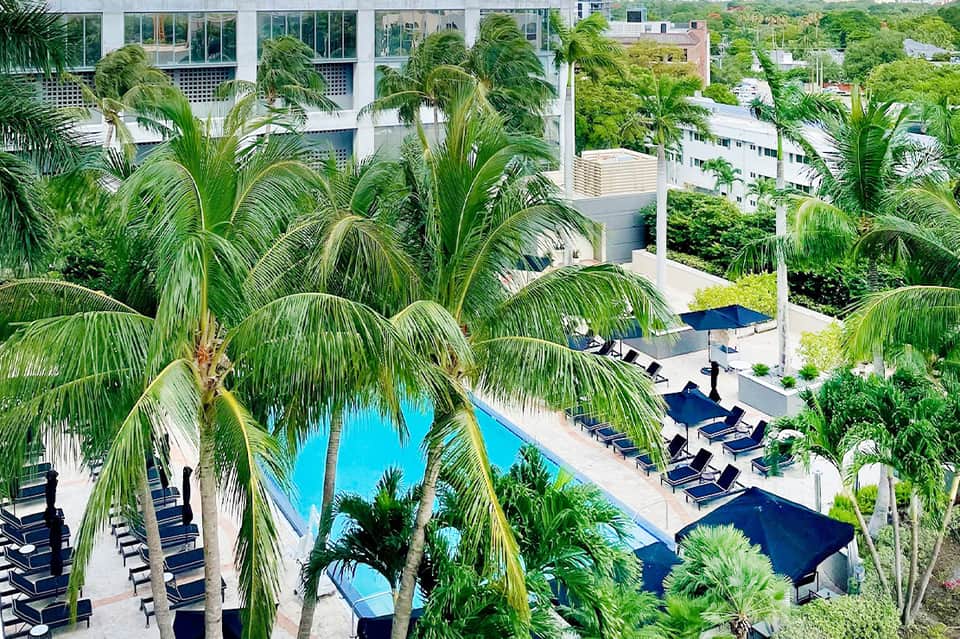 Aerial view of resort pool surrounded by palm trees, lounge chairs with blue umbrellas, and modern white building architecture