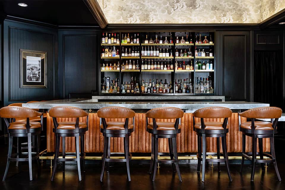 Upscale bar with illuminated shelving displaying spirits, leather bar stools, and marble countertop