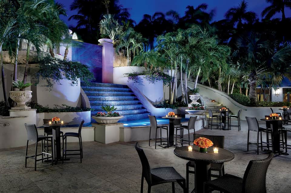 Illuminated courtyard at night with blue-lit staircase, lounge seating, palm trees, and water features