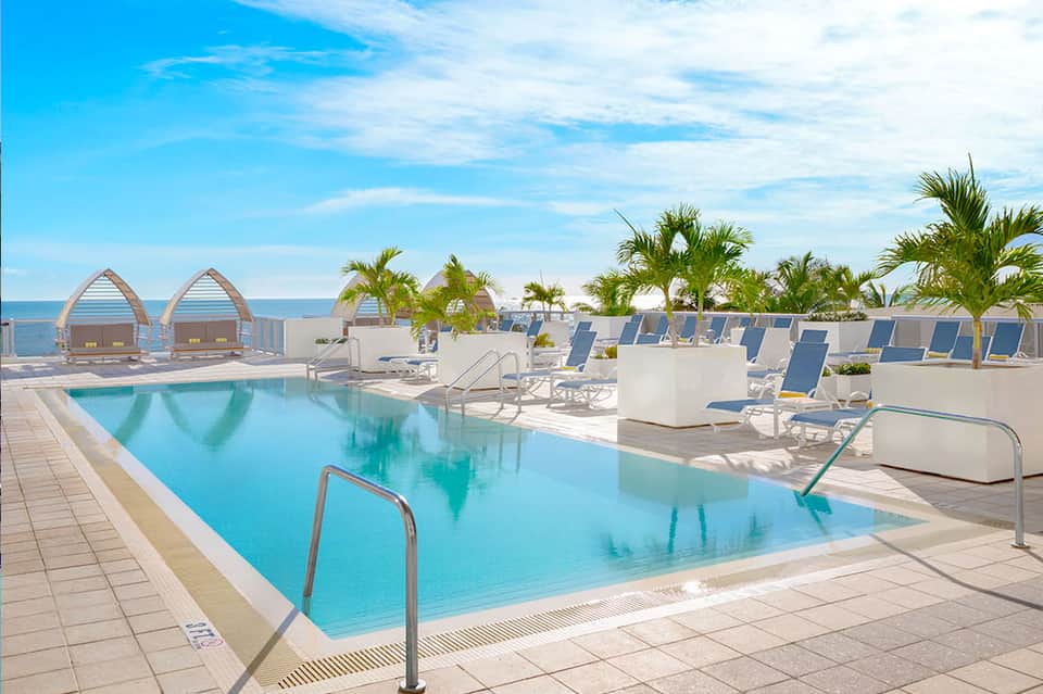Beachfront resort pool with blue lounge chairs, palm trees, and ocean view cabanas