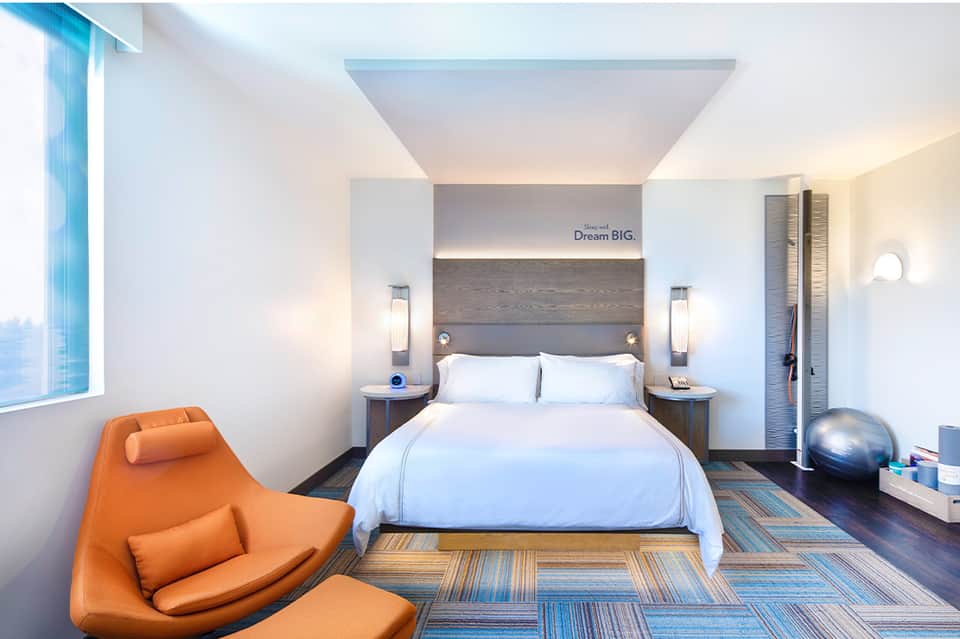 Modern hotel bedroom with white bedding, orange accent chair, and contemporary design elements