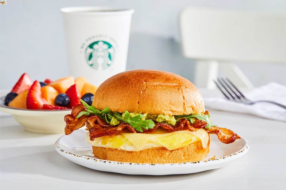 Breakfast plate with bacon and egg sandwich, fresh fruit, and Starbucks coffee on white table setting.