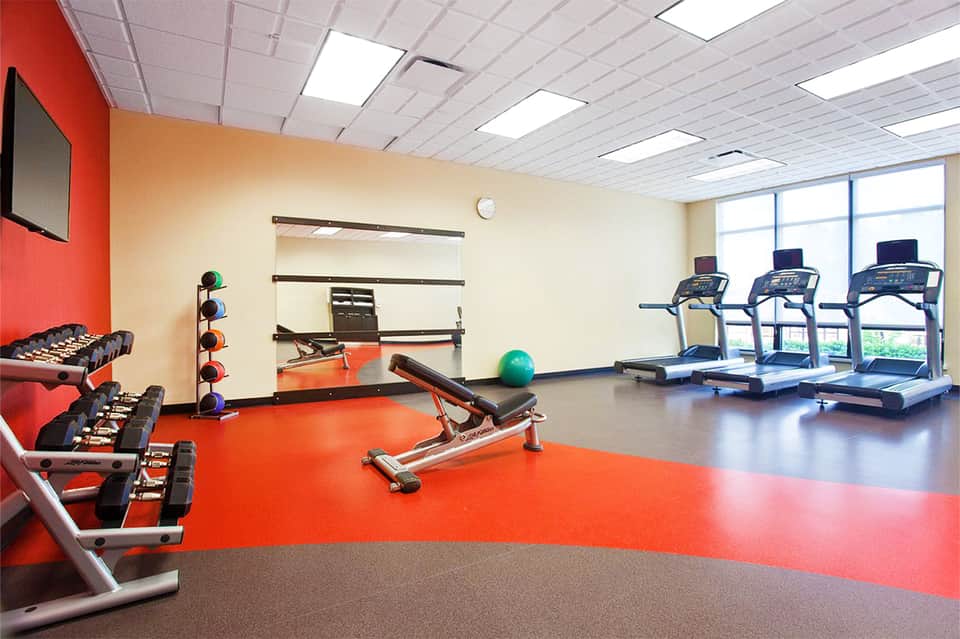 Modern fitness center with red and gray flooring, dumbbells, treadmills, and weight training equipment