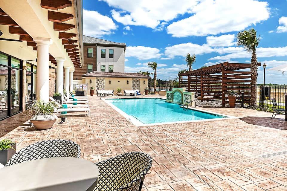 Resort pool deck with lounge chairs, pergola shade structures, and main building with arches