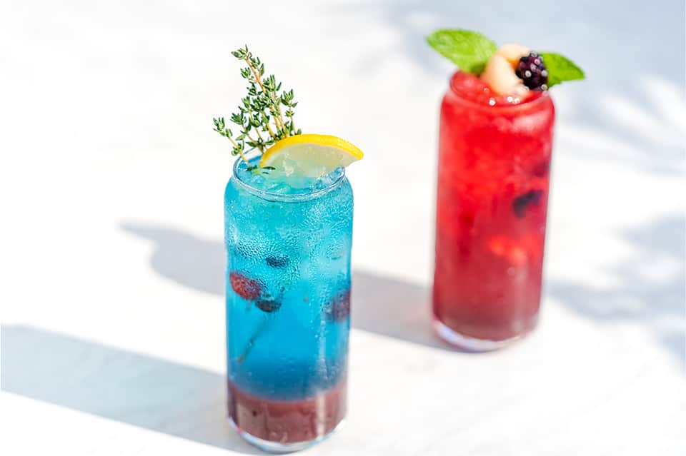Two colorful tropical cocktails with fresh fruit garnish on white surface in bright sunlight