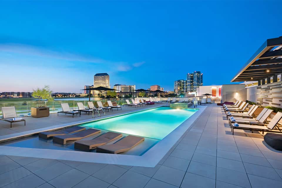 Modern rooftop pool at dusk with illuminated water, lounge chairs, and city skyline in background